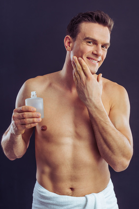 Handsome naked man is smiling, holding aftershave lotion and applying it, on a dark background